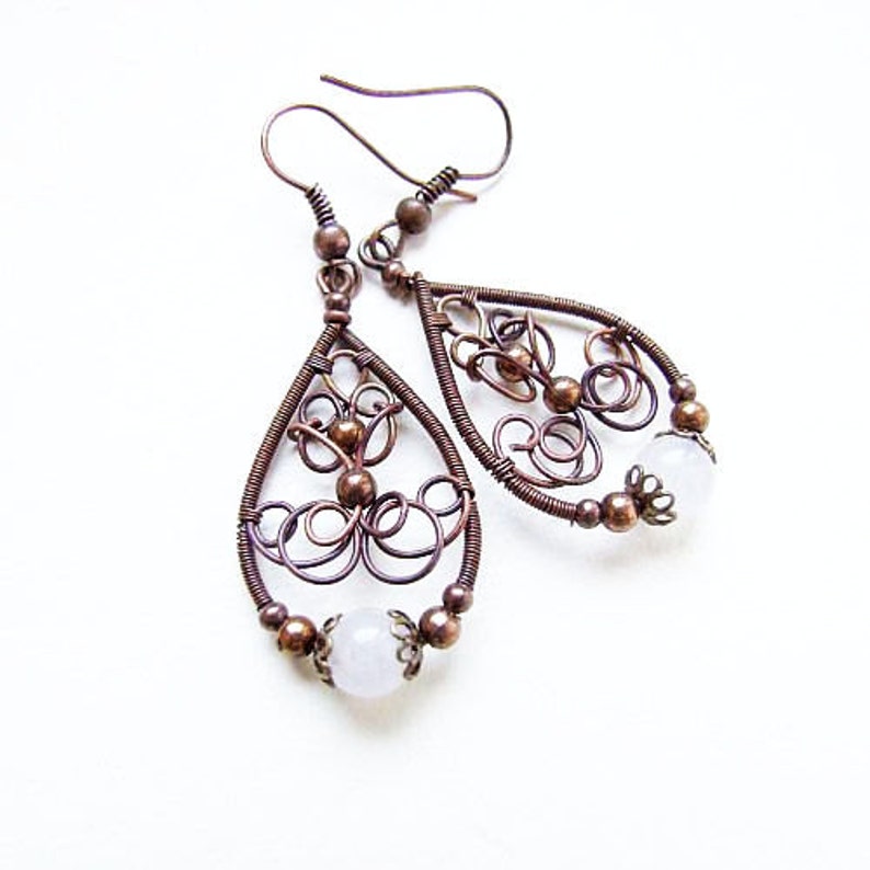 May include: A pair of copper wire earrings with a delicate, intricate design. Each earring features a teardrop shape with a white stone at the bottom.
