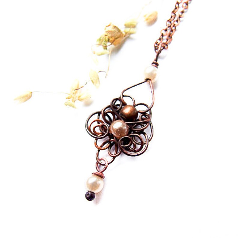 May include: A copper wire necklace with a delicate, intricate design featuring a cluster of brown and white pearls. The necklace is suspended from a simple chain.