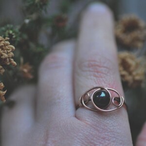Puede incluir: Un anillo de alambre color cobre con una piedra oscura y redonda en el centro. El anillo est&aacute; en un dedo, con flores secas en el fondo. El dise&ntilde;o del anillo presenta un delicado patr&oacute;n arremolinado alrededor de la piedra.