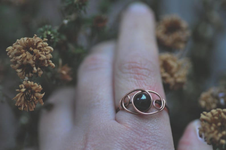 Puede incluir: Un anillo de alambre color cobre con una piedra oscura y redonda en el centro. El anillo est&aacute; en un dedo, con flores secas en el fondo. El dise&ntilde;o del anillo presenta un delicado patr&oacute;n arremolinado alrededor de la piedra.
