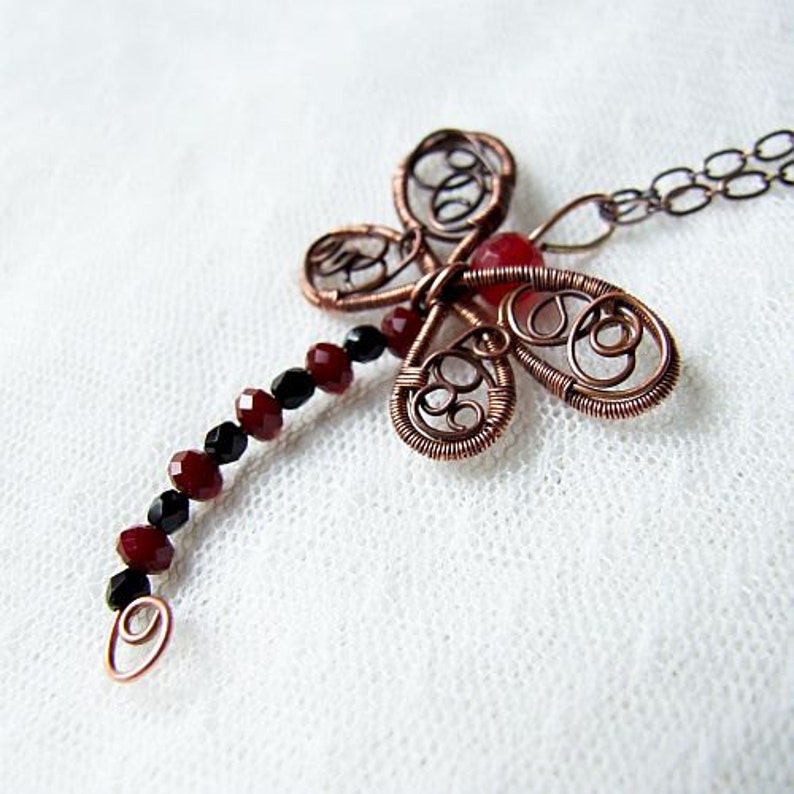 Pu&ograve; includere: Una collana con un ciondolo a forma di libellula in filo di rame con perline rosse e nere. La libellula ha un lavoro di filo intricato e una pietra preziosa rossa al centro.