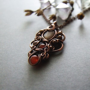 Könnte beinhalten: Ein mit Kupferdraht umwickelter Anhänger mit drei Steinen. Die Steine sind hellbraun, dunkelrot und hellrot. Der Anhänger hängt an einer Kupferkette.