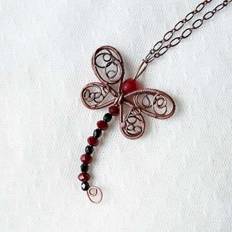 Pu&ograve; includere: Una collana con un ciondolo a forma di libellula in filo di rame con perline nere e rosse. La libellula ha un lavoro di filo intricato e una pietra preziosa rossa al centro.