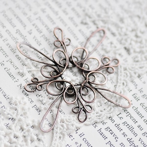 Pode incluir: Pingente artesanal em arame de cobre com um design simétrico em forma de flor. A peça intrincada apresenta elementos de arame em laço e enrolados, criando um padrão vazado. O pingente é exibido em um fundo de papel vintage.