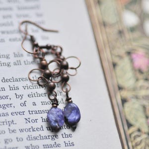 May include: A pair of earrings with copper wire and purple gemstone beads. The earrings are hanging from a book.