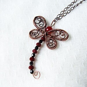 Pu&ograve; includere: Una collana con un ciondolo a forma di libellula in filo di rame, una pietra preziosa rossa e perline nere. La libellula presenta dettagli intricati in filo di ferro.