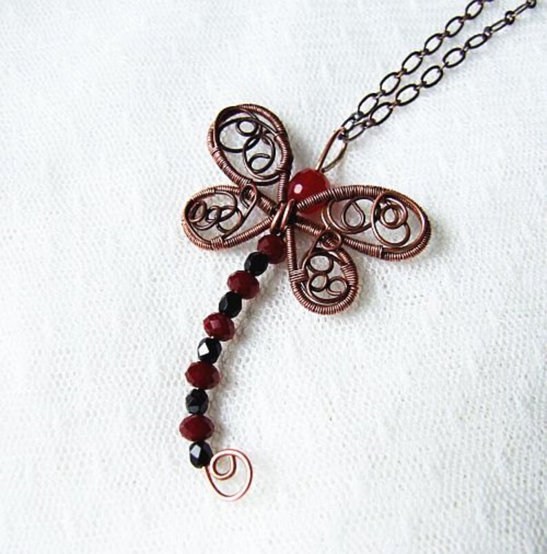 Pu&ograve; includere: Una collana con un ciondolo a forma di libellula in filo di rame, una pietra preziosa rossa e perline nere. La libellula presenta dettagli intricati in filo di ferro.