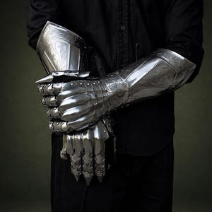 May include: A pair of polished steel gauntlets featuring intricate etched scrollwork patterns. The armor pieces cover the hands and forearms, with articulated finger plates and pointed metal tips, showcasing a reflective metallic finish.
