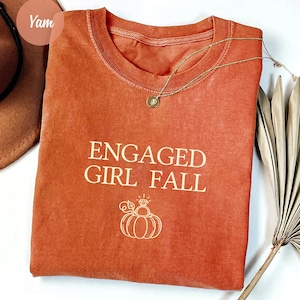 May include: A rust-colored short-sleeve t-shirt featuring the text ENGAGED GIRL FALL in cream-colored lettering. Below the text is a line-art graphic of a pumpkin topped with a diamond engagement ring. A gold-toned necklace rests on the neckline.