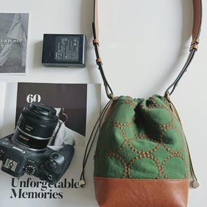 May include: A green bucket bag with a brown leather base and a brown stitched circular pattern. The bag features a drawstring closure and a long, adjustable brown shoulder strap. The bag is positioned next to a black camera and a battery charger.