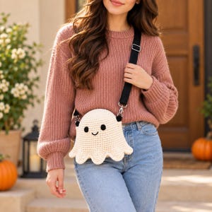 Può includere: Una borsa a tracolla all'uncinetto color crema a forma di fantasma con occhi ricamati neri e un piccolo sorriso. La borsa presenta una spessa tracolla nera regolabile con finiture in metallo argentato, indossata sopra un maglione in maglia rosa.