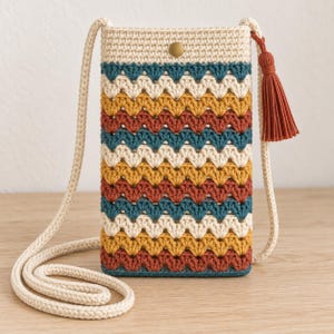 May include: A rectangular crochet crossbody bag featuring a chevron pattern in alternating stripes of cream, mustard yellow, terracotta, and teal. The bag has a cream-colored strap, a single brass-tone snap closure, and a terracotta-colored tassel.