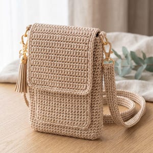 Op de afbeelding: Een beige gehaakte schoudertas met een rechthoekige klepsluiting, een bijpassende lange schouderriem en een decoratieve kwast bevestigd aan een goudkleurige metalen sluiting aan de zijkant. De tas is gemaakt met een dicht, gestructureerd patroon.