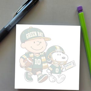 May include: A square notepad featuring a cartoon illustration of a boy wearing a Green Bay cap and jersey with the number 10, holding a football, and a dog wearing a football helmet and jersey with the number 0, holding a Game Day ticket.