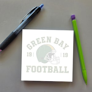 May include: A square white sticky note featuring a graphic of a football helmet in the center, with the text "GREEN BAY" above, "FOOTBALL" below, and "19" on both sides. A black retractable pen and a lime green pencil are positioned nearby.