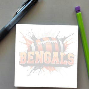 May include: A square white sticky note featuring a graphic of an American football with orange and black stripes, surrounded by a black and orange paint splatter effect, with the word BENGALS printed in large orange block letters.