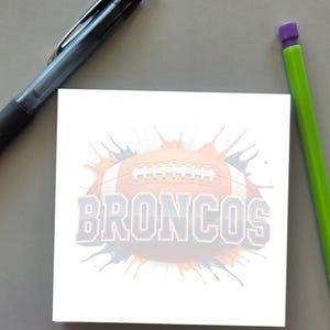 May include: A square white sticky note featuring a graphic of a brown football with white laces, surrounded by orange and blue paint splatters, and the word BRONCOS printed in large, bold, light blue block letters with a dark outline.