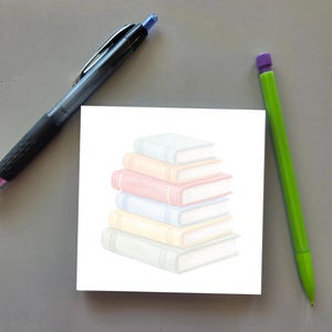 May include: A square white sticky note featuring a central illustration of a stack of six books in pastel shades of blue, peach, pink, light blue, yellow, and gray, accompanied by a black retractable pen and a bright green pencil.