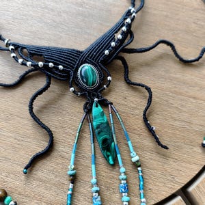 May include: A black macrame necklace featuring a central oval malachite cabochon set in silver, with a dangling malachite shard and multiple strands of turquoise, blue, and silver beads extending downwards from the main woven structure.