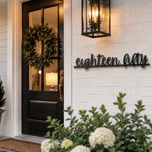 May include: A black metal wall sign featuring the cursive text eighteen fifty mounted on a white brick wall next to a black front door with a green wreath, a black outdoor lantern, and a potted plant in the foreground.