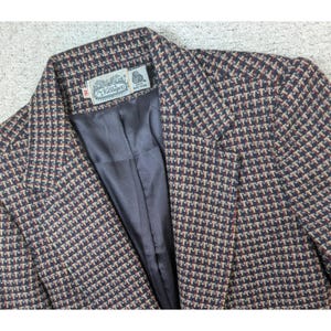 May include: A blazer featuring a multi-colored houndstooth pattern in shades of navy blue, red, and tan. The interior is lined with a solid dark gray fabric. A white label inside the collar reads The Villager, 100% Pure Wool, and the size 16.