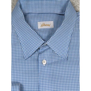 May include: A light blue and white gingham button-down shirt featuring a pointed collar and a single visible white button. A white rectangular label with the gold text Brioni is stitched onto the inside of the collar area of the garment.