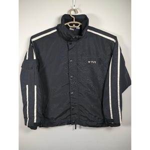 May include: A black jacket with a high collar, featuring white vertical stripes running down the length of each sleeve. The front includes a central snap-button closure, a small zippered chest pocket with the TYR logo, and side pockets.