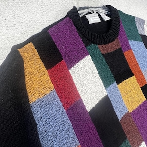 Puede incluir: Un suéter de manga larga con un patrón de bloques multicolor que incluye cuadrados en negro, morado, amarillo mostaza, rojo, blanco, verde oscuro, azul claro y marrón. La prenda tiene un cuello redondo acanalado negro y una etiqueta blanca.