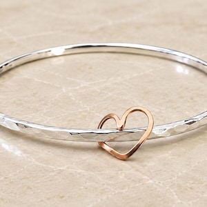Puede incluir: Una pulsera rígida de plata con una textura martillada y reflectante. La banda circular presenta un dije de corazón de contorno abierto hecho de metal de color rosa que está enhebrado en la pulsera, creando un contraste de dos tonos.