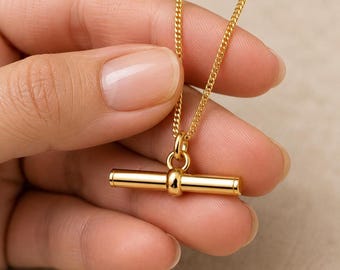 May include: A gold-toned necklace featuring a polished cylindrical bar pendant suspended from a delicate curb chain. The pendant is attached to the chain by a small circular jump ring, creating a clean, metallic aesthetic with a smooth finish.