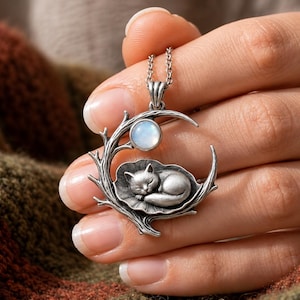 May include: A silver-toned pendant necklace featuring a sleeping cat curled on a leaf, nestled within a crescent moon frame with branch-like details and a small, round, iridescent white stone set at the top, suspended from a silver chain.
