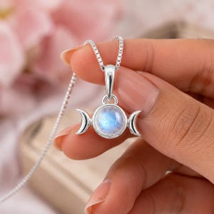 May include: A silver necklace featuring a circular pendant with a central iridescent moonstone cabochon flanked by two silver crescent moon shapes. The pendant hangs from a delicate silver box chain, showcasing a polished metallic finish.