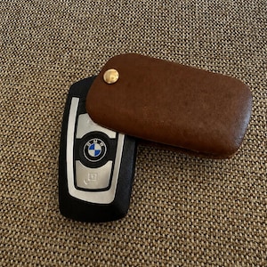 May include: A black BMW car key fob partially covered by a brown leather protective case. The key fob features the blue and white BMW logo in the centre, silver-toned accents, and a gold-coloured metal snap button on the leather cover.