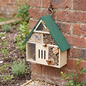 May include: A wooden insect hotel with a dark green gabled roof, featuring multiple compartments filled with bamboo tubes, wood shavings, and pinecones, protected by wire mesh, with a central wooden panel and two butterflies resting on it.