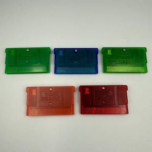 May include: Five rectangular video game cartridges with translucent plastic shells. The collection includes two green, one blue, and two red cartridges. Each unit features a metallic screw, a connector edge, and embossed Nintendo branding text.