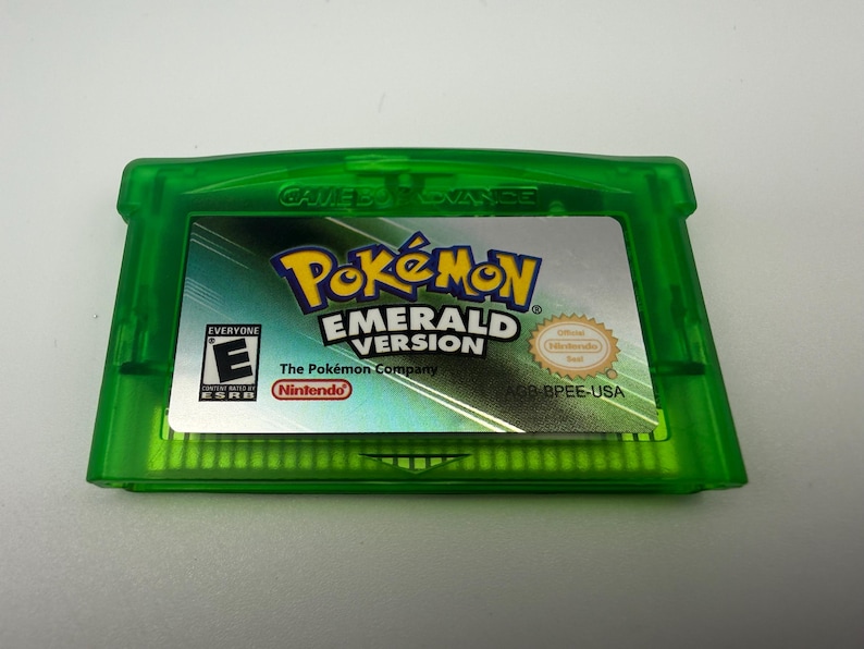 May include: A translucent green Game Boy Advance video game cartridge for Pok&eacute;mon Emerald Version. The label features the game title, the ESRB E rating, the Nintendo logo, and the text AGB BPEE-USA printed on a silver background.