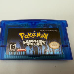 May include: A translucent blue Game Boy Advance video game cartridge for Pok&eacute;mon Sapphire Version. The label features the Pok&eacute;mon logo, the text Sapphire Version, an ESRB E rating, the Nintendo logo, and the official Nintendo seal.