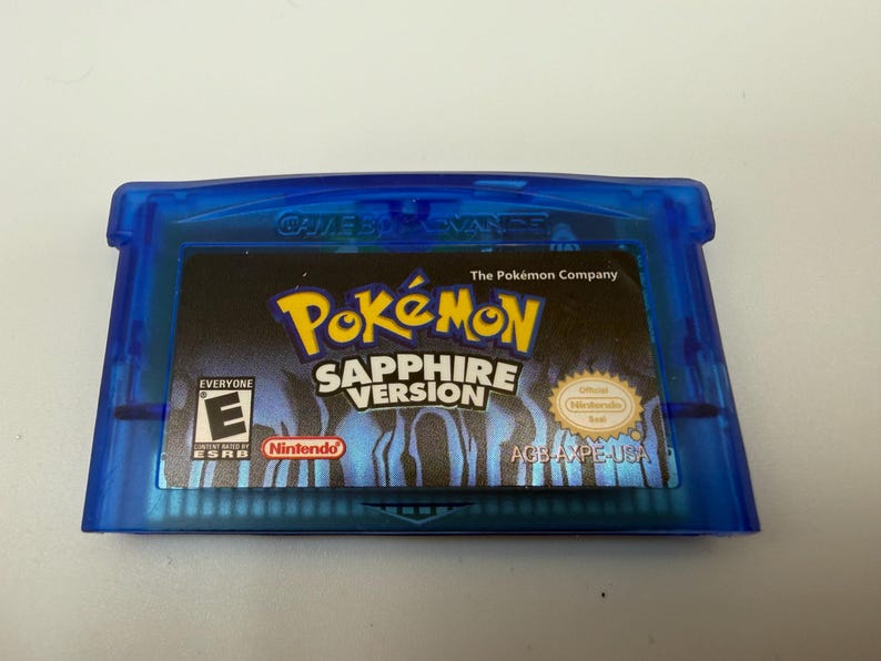 May include: A translucent blue Game Boy Advance video game cartridge for Pok&eacute;mon Sapphire Version. The label features the Pok&eacute;mon logo, the text Sapphire Version, an ESRB E rating, the Nintendo logo, and the official Nintendo seal.