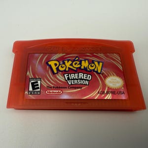 May include: A translucent red Game Boy Advance video game cartridge for Pok&eacute;mon FireRed Version. The label features the Pok&eacute;mon logo, the text FireRed Version, an ESRB E rating, the Nintendo logo, and the product code AGB-BPRE-USA.
