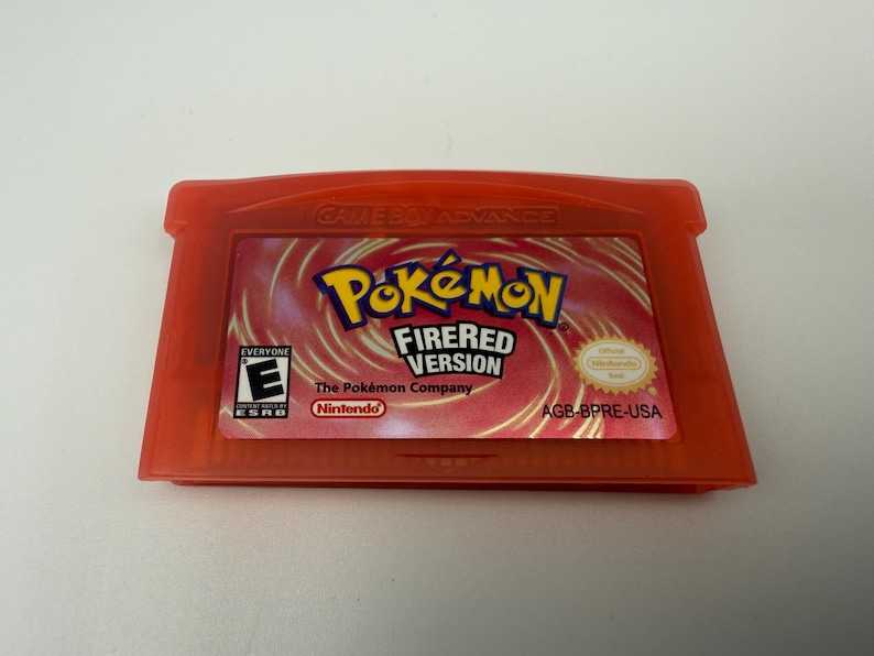 May include: A translucent red Game Boy Advance video game cartridge for Pok&eacute;mon FireRed Version. The label features the Pok&eacute;mon logo, the text FireRed Version, an ESRB E rating, the Nintendo logo, and the product code AGB-BPRE-USA.