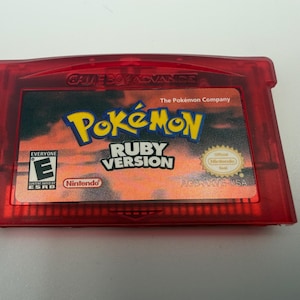 May include: A translucent red Game Boy Advance video game cartridge for Pok&eacute;mon Ruby Version. The label features a sunset-colored background, the Pok&eacute;mon logo, the ESRB Everyone E rating, and the official Nintendo Seal of Quality.