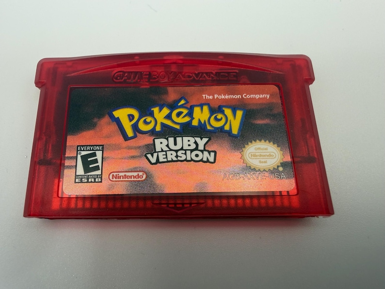 May include: A translucent red Game Boy Advance video game cartridge for Pok&eacute;mon Ruby Version. The label features a sunset-colored background, the Pok&eacute;mon logo, the ESRB Everyone E rating, and the official Nintendo Seal of Quality.