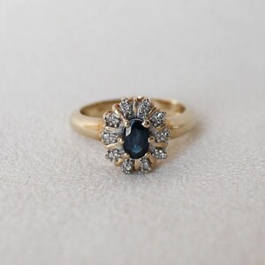 Può includere: Un anello in oro con uno zaffiro blu scuro centrale a taglio ovale, circondato da un alone di piccoli diamanti bianchi rotondi e chiari. La montatura presenta un design a grappolo floreale, con le pietre incastonate su una fascia in oro.