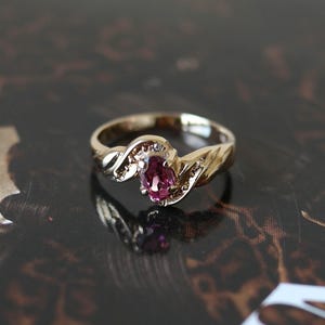 Op de afbeelding: Een gouden ring met een centrale ovaal geslepen roze edelsteen in een klauwzetting. De band heeft een gepolijste afwerking en bevat kleine, heldere accentstenen ingebed langs de zijkanten van de zetting, in een overlappend ontwerp.