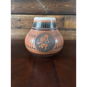 May include: A terracotta-colored ceramic vase with a rounded body and a narrow neck. The exterior features a central circular medallion depicting a stylized figure playing a flute, surrounded by etched geometric patterns in black and dark brown.