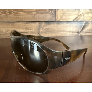 May include: Tortoiseshell-patterned sunglasses featuring a wrap-around frame design with dark tinted lenses. The word POLO is printed in white on the outer temple arm, and the brand name is also visible on the inner temple arm of the eyewear.