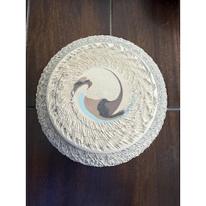 May include: A round ceramic plate with a textured, light gray rim featuring a radial pattern. The center is smooth and off-white, decorated with a swirling abstract design in shades of brown, black, and light blue, creating a dynamic visual effect.