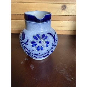 Puede incluir: Una jarra de cerámica redonda con un acabado de esmalte de sal gris claro texturizado, un borde azul cobalto profundo y un patrón floral prominente en el mismo tono azul. La jarra tiene un cuerpo ancho y bulboso con un pequeño pico vertedor.