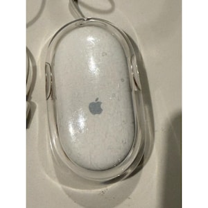 May include: A white computer mouse with a clear, transparent outer shell casing. The top surface of the mouse features a centered, grey Apple logo. The device has an oval shape and includes a white cord extending from the top edge of the unit.