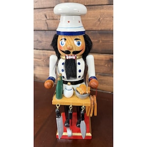 May include: A wooden nutcracker figurine dressed as a chef, wearing a white hat with red accents and a white jacket with blue buttons. The figure holds a small wooden tray containing a green bottle, a white shaker, and a bowl with food items.